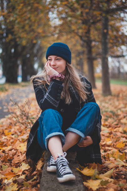 Smiling and happy brunette sitting in a pile of colourful leaves in a city park. Candid portrait of a young real woman in autumn clothes in the fall season. Orange, red and brown colour. - Foto, Imagen