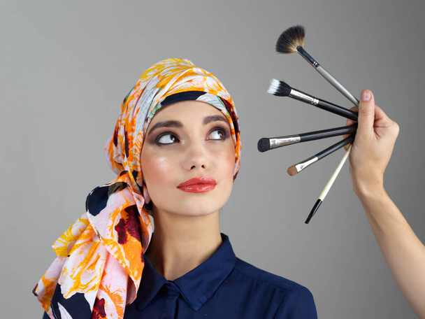 May you be as flawless as your makeup too. Studio shot of a confident young woman wearing a colorful head scarf while looking at different makeup brushes being held in front of her - Foto, imagen