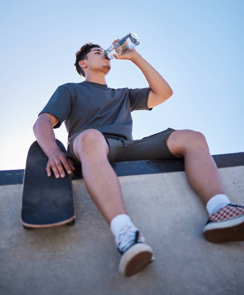 Skateboard, young man and drinking water bottle, relax and on break on sunny day outdoor. Male, skater and athlete hydrate, thirsty and sports for wellness, health and have fun in summer - Photo, image