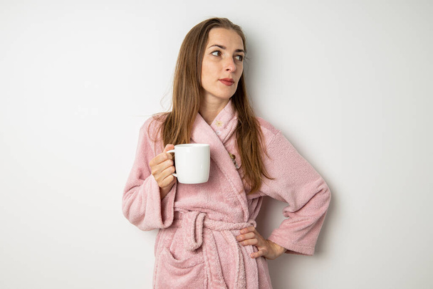 Νεαρή γυναίκα με μπουρνούζι πίνει καφέ σε λευκό φόντο. - Φωτογραφία, εικόνα