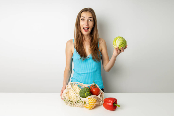 Verrast jonge vrouw nemen van groenten uit zak op tafel op witte achtergrond. - Foto, afbeelding