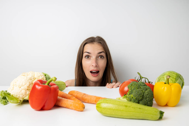 Fiatal nő kukucskál ki az asztal alól meglepetten nézi a friss zöldségeket.. - Fotó, kép