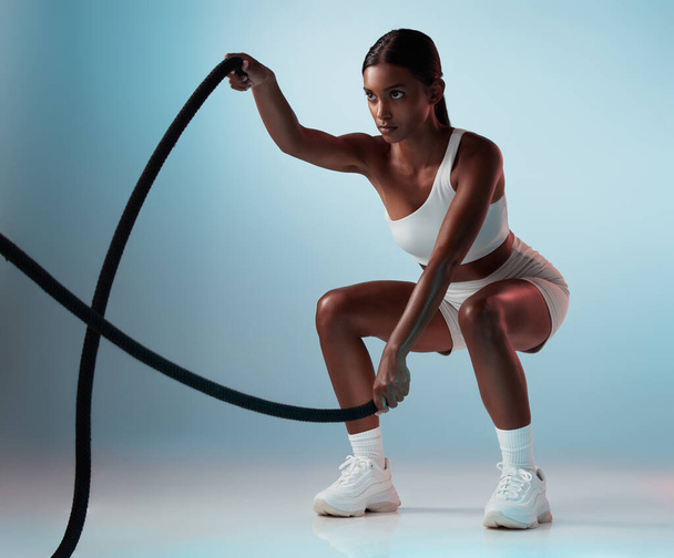 Fitness, woman and rope exercise, sports training and workout for muscle, energy and wellness on blue background. Focus indian girl athlete moving battle ropes with speed, action and power in studio. - Fotografie, Obrázek