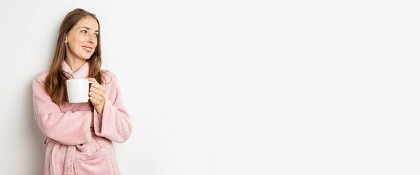 Smiling young woman in a bathrobe with a cup on a white background. Banner. - Photo, Image