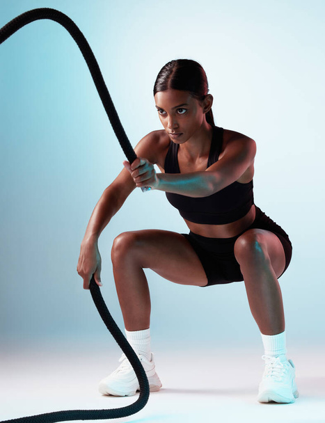 Battle rope, exercise and fitness model in a studio with energy, motivation and training. Healthy, strong and slim woman athlete from India doing a cardio sports workout isolated by a blue background. - Fotoğraf, Görsel