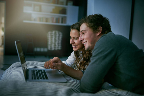 Tu n'as pas besoin de sortir pour sortir ce soir. un jeune couple utilisant leur ordinateur portable ensemble tout en étant couché sur le lit - Photo, image