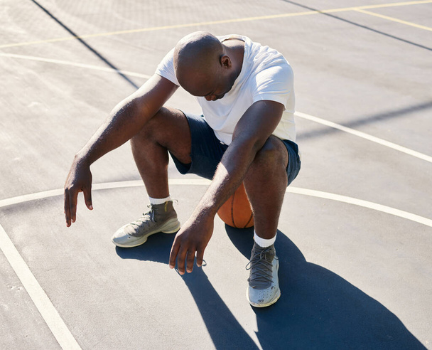 Basketball court, tired and black man sitting on a basketball resting or relaxing on a fitness training or workout break. Loser, wellness and African sports athlete with fatigue in an exercise game. - Photo, Image