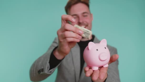 Gelukkige jonge zakenman ondernemer geld besparen met behulp van roze piggybank zetten een dollar gegoten in. Succes, economie, investeringen, storting. baard jongen geïsoleerd alleen op blauw studio achtergrond - Video