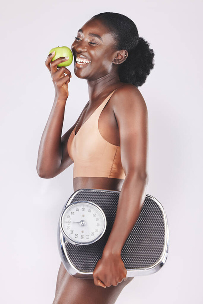 Mulher feliz, escala e maçã para perder peso, dieta saudável e resultados de fitness, progresso e motivação de exercício no fundo do estúdio. Modelo preto comer frutas, nutrição esportiva e bem-estar corporal. - Foto, Imagem