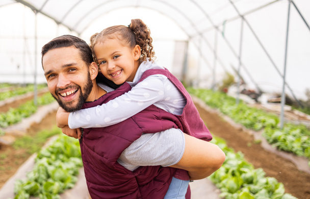 Greenhouse farmer, dad with girl and happy smile of girl getting piggyback ride from father in an organic farm for sustainable crop growth. Healthy food produce, natural crops and modern agriculture. - Foto, afbeelding