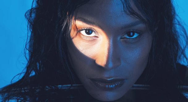 Beauty, cosmetics and portrait of a model with a light reflection on her face in the studio. Spotlight, skincare and young woman from Puerto Rico with natural skin isolated by a blue background - Fotoğraf, Görsel