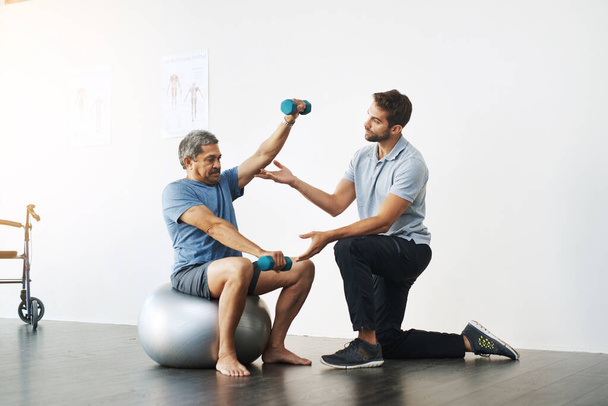 Αργές και εύκολες κινήσεις. ένας νεαρός άνδρας φυσιοθεραπευτής που βοηθά έναν ώριμο άνδρα ασθενή με κινητικές ασκήσεις σε μια κλινική - Φωτογραφία, εικόνα