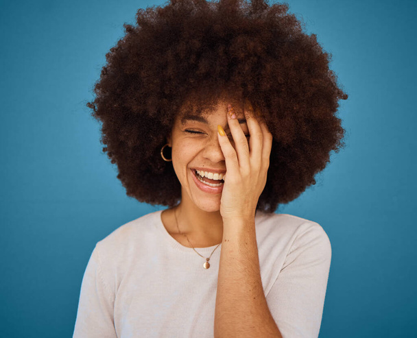 Face, funny and laughing with an afro black woman in studio on a blue background for fun or humor. Smile, comic and happy with an attractive young female feeling positive with joy or comedy. - Foto, Imagem