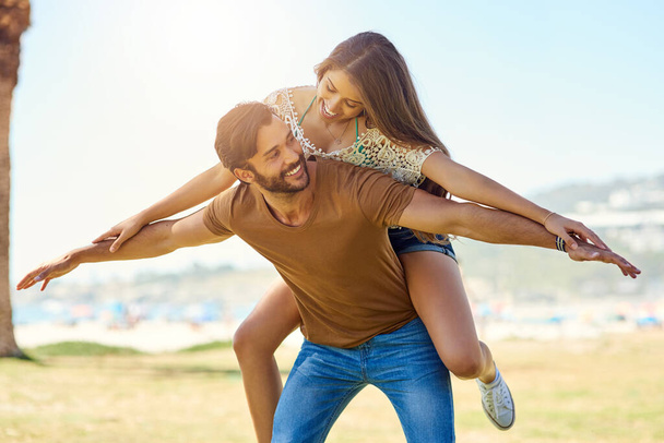 Liefde tilt ons op. een glimlachende jonge man het geven van zijn vriendin een piggyback terwijl genieten van een dag samen buiten - Foto, afbeelding