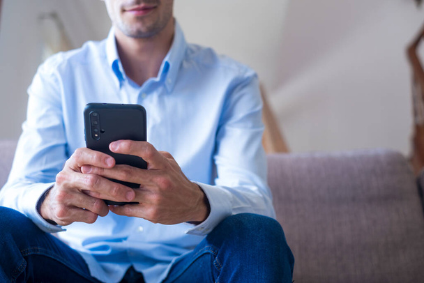 Hands of man sitting on sofa and text messaging using mobile phone at home. Caucasian guy browsing social media content or chatting online on smartphone while relaxing on couch   - Фото, изображение