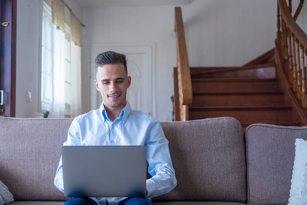 Mladý pohledný usměvavý chlapík si prohlíží laptop, sedí doma na útulné pohovce v obývacím pokoji. Šťastný běloch pracující online nebo sledující obsah sociálních médií na gauči v moderním bytě  - Fotografie, Obrázek