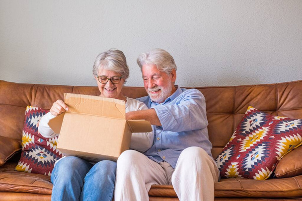 Старая пара распаковывает коробку с доставкой дома. Счастливая пожилая пара смотрит на картонную коробку, сидя дома на диване в гостиной. Пожилая пара проверяет доставленные вещи дома.  - Фото, изображение