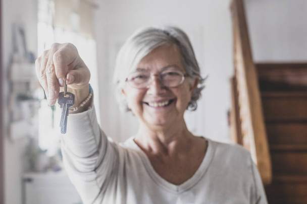 Egy boldog idős nő portréja, kezében az új ház vagy tulajdon kulcsaival. Öreg hölgy bemutatja vagy odaadja a ház kulcsait. Idős nő boldog, hogy új lakásba költözik  - Fotó, kép