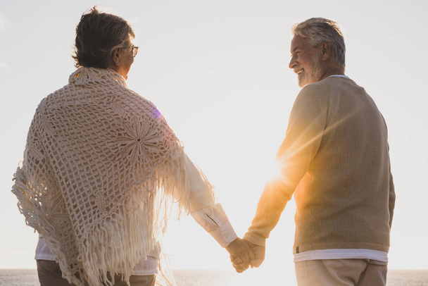 pareja de dos personas mayores felices y activas divirtiéndose y disfrutando juntos del verano en la playa caminando tomados de la mano con la puesta de sol al fondo  - Foto, imagen
