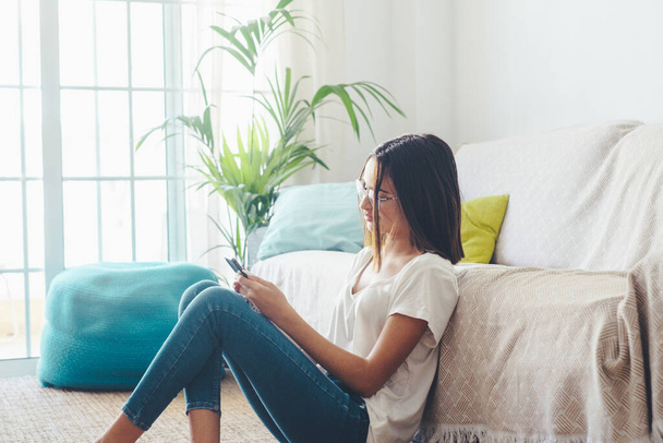Σοβαρή γυναίκα με γυαλιά ηλίου γραπτό μήνυμα χρησιμοποιώντας κινητό τηλέφωνο, ενώ κάθεται στο πάτωμα στο σαλόνι του σπιτιού της. Όμορφη κυρία ξοδεύει ελεύθερο χρόνο χρησιμοποιώντας το smartphone στο σπίτι  - Φωτογραφία, εικόνα