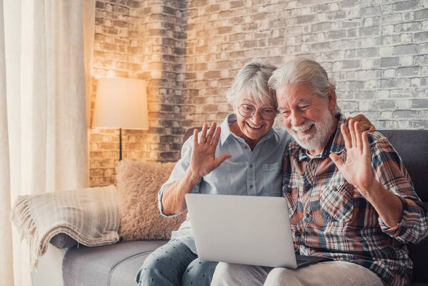 Cute couple of old people sitting on the sofa using laptop together shopping and surfing the net. Two mature people in the living room enjoying technology talking in video call with friends or family.  - Foto, Imagen