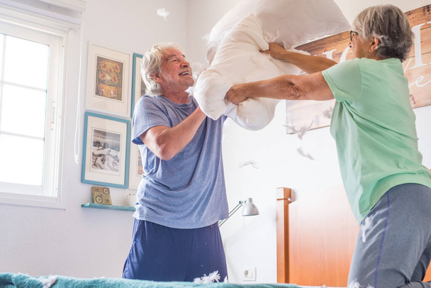 ζευγάρι των δύο ευτυχισμένων ηλικιωμένων που διασκεδάζουν παίζοντας μαζί στο κρεβάτι στο σπίτι αγωνίζονται με μαξιλάρια απολαμβάνοντας - μαξιλάρια πολέμου σε εσωτερικούς χώρους το πρωί  - Φωτογραφία, εικόνα