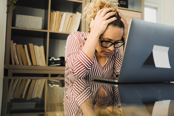 Jedna samotna, nieszczęśliwa i smutna kobieta w biurze, używająca laptopa i patrząca na niego z rękami w głowie. Zestresowana kobieta w tarapatach rozwiązuje problem  - Zdjęcie, obraz