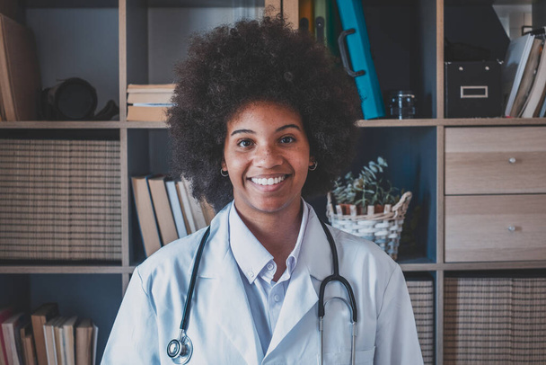 Médica afro-americana feliz com estetoscópio na clínica. Trabalhador de saúde da linha de frente confiante sorrindo enquanto olha para a câmera. Jovem médico vestindo jaleco com estetoscópio no escritório do hospital   - Foto, Imagem