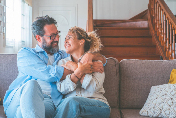 Happy caucasian couple looking at each other sitting on sofa in the living room. Loving husband and wife embracing each other sitting on couch at home. Man and woman spending leisure time at modern apartment - Photo, Image