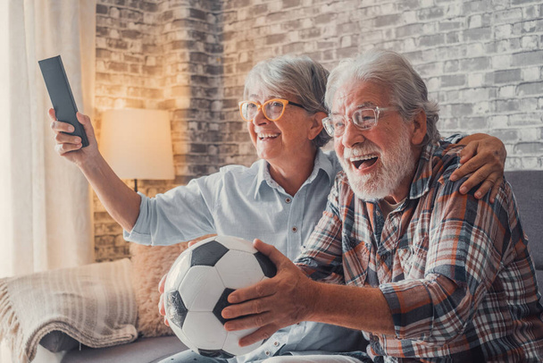 Ευτυχισμένο ζευγάρι ηλικιωμένων ή οπαδών ποδοσφαίρου βλέποντας ποδόσφαιρο στην τηλεόραση και γιορτάζοντας τη νίκη στο σπίτι. Οικογενειακή έννοια, έννοια αθλητισμού και ψυχαγωγίας. - Φωτογραφία, εικόνα