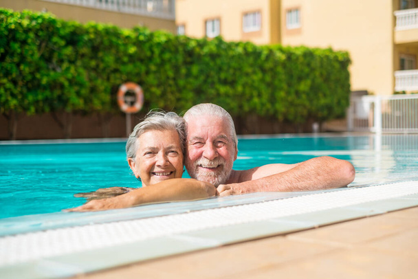 Pareja de dos felices seniors divirtiéndose y disfrutando juntos en la piscina sonriendo y mirando a la cámara. Gente feliz disfrutando del verano al aire libre en el agua  - Foto, Imagen