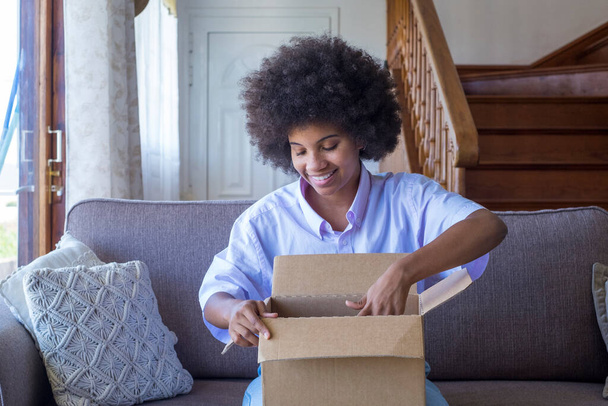 Африканка сидить на дивані в вітальні, тримаючись за велику коробку з картонними коробками, яку розбирає онлайн-магазин. Цікава чорна леді розпаковує картонні пакунки. Усмішка самка без боксу - Фото, зображення