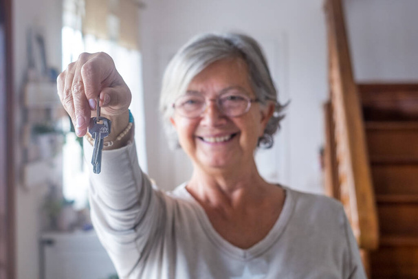 Close-up van oude en volwassen vrouw thuis met huissleutels van de camera. Portret van vrouwelijke senior lachend en kijkend naar de camera.  - Foto, afbeelding