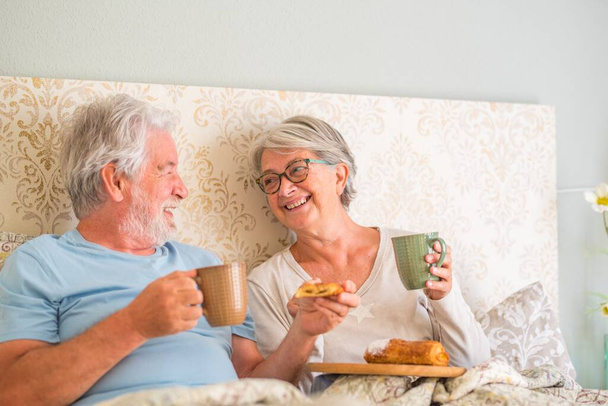 Старая старшая кавказская пара смеется и наслаждается завтраком в постели в спальне дома. Пожилая пара ест круассан и пьет кофе из чашки на завтрак дома.  - Фото, изображение