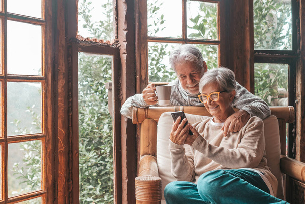 Couple of two old and mature people at home using phone together in sofa. Senior use smartphone having fun and enjoying looking at it. Leisure and free time concept in the living room  - Photo, Image