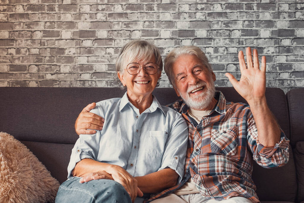 Χαριτωμένο και όμορφο ζευγάρι ηλικιωμένων που χαμογελούν και κοιτάζουν την κάμερα και μιλάνε στο σπίτι μαζί. Πορτρέτο των ηλικιωμένων που κάθονται στον καναπέ απολαμβάνοντας και χαλαρώνοντας και χαιρετώντας με το χέρι.  - Φωτογραφία, εικόνα