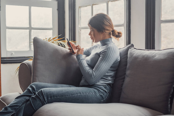 Nuori kaunis nainen istuu sohvalla kotona juttelemassa ja surffaamassa netissä. Naishenkilö hauskaa älypuhelimen verkossa. Muotokuva tyttö hymyilee käyttäen matkapuhelin tarkistaa sosiaalisen median  - Valokuva, kuva