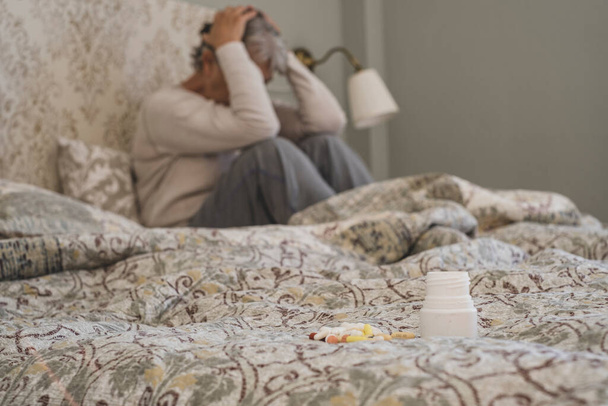 Tablety nebo pilulky na posteli se starší ženou trpící depresí, která sedí v pozadí. Stará dáma drží hlavu v silné bolesti migrény, neklid a nepříjemné v ložnici s léky v popředí   - Fotografie, Obrázek