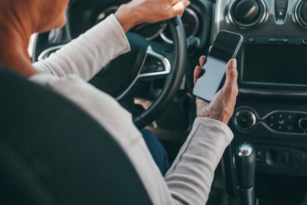 Portret van een oude vrouw die een telefoon in de auto gebruikt tijdens het rijden op de weg, kan een ongeluk veroorzaken. Close-up van handen houden smartphone surfen online sms 'en en chatten.  - Foto, afbeelding