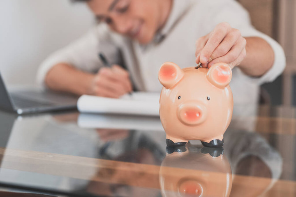 Man sit at desk manage expenses, calculate expenditures, pay bills online use laptop, makes household finances analysis, close up focus on pink piggy bank. Save money for future, be provident concept  - Photo, Image