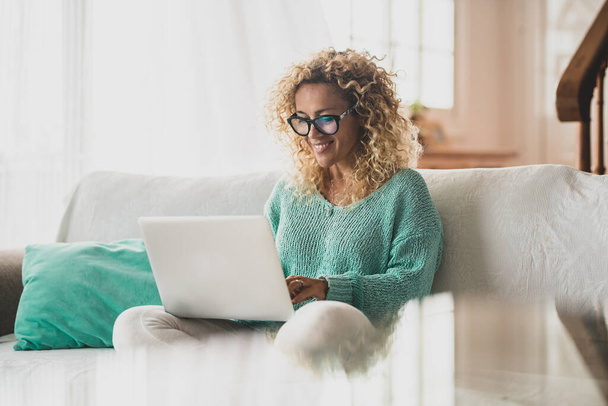 одна счастливая кудрявая женщина, сидящая на диване своего дома, используя компьютер для серфинга в сети - красивая дама, использующая ноутбук на диване своего дома  - Фото, изображение