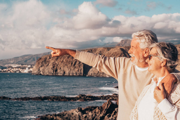 Ritratto di coppia di maturi e anziani che si godono l'estate in spiaggia guardando il mare sorridente e divertendosi insieme al tramonto sullo sfondo. Due anziani attivi che viaggiano all'aperto.  - Foto, immagini