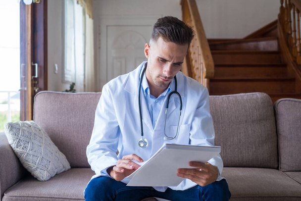 Focused young male doctor in the sofa and medical coat working with paper registry book, making notes, managing patients visits, recording healthcare treatment, sitting on couch at home.   - Photo, Image