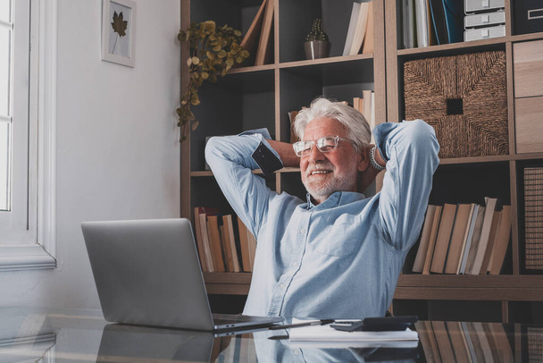 Happy satisfied senior businnessman relaxing with his hands behind head and eyes closed at home office. Dreamy old man resting at workplace, feeling peace of mind after finishing his work - Foto, immagini