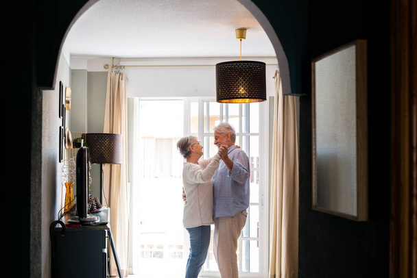 Romántica pareja de ancianos amantes tomados de la mano disfrutando bailando juntos en la sala de estar de la casa, pareja abrazándose mientras se toman de la mano juntos en casa  - Foto, imagen