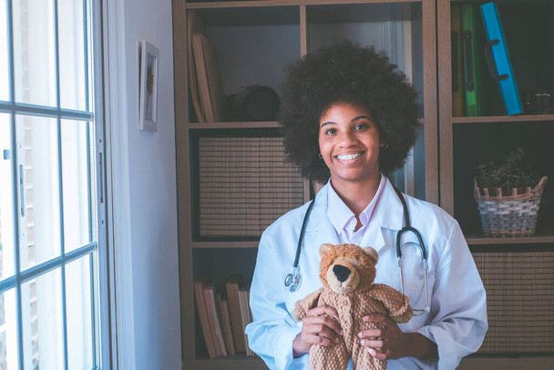 Portrait of young african american female nurse in medical uniform holding stuffed toy showing care love to little patient, happy woman doctor pediatrician posing with teddy bear in hospital or clinic - Foto, imagen
