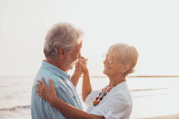 Couple of old mature people dancing together and having fun on the sand at the beach enjoying and living the moment. Portrait of seniors in love looking each others having fun.  - Photo, Image