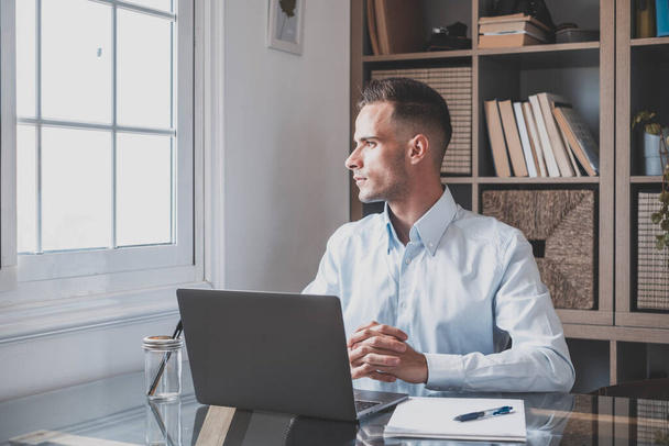 Serious young businessman sitting at office and looking out through window thinking about future career or success. Thoughtful caucasian guy sitting at desk with laptop while looking away and admiring view  - Photo, Image