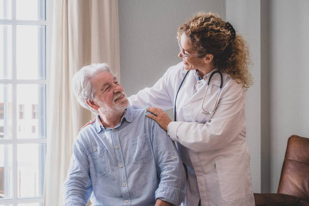 Young female doctor encouraging senior man at home. Doctor helping senior patient and giving care. Female home caregiver taking care of retired old man. Old man and physician talking at nursing home - Photo, Image