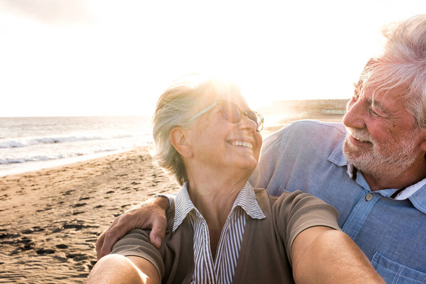 Portret pary dojrzałych i starszych ludzi cieszących się latem na plaży patrzących na aparat fotograficzny robiący sobie selfie wraz z zachodem słońca w tle. Dwóch aktywnych seniorów podróżujących na zewnątrz.  - Zdjęcie, obraz